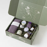 A lavender-themed gift box set shown from an overhead angle, featuring a sage green box with white botanical illustrations, filled with lavender-scented items including a purple towel, small jars, dried lavender, and lavender mist on a white background.