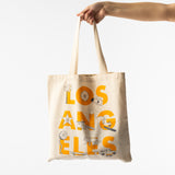 A hand displays the Los Angeles Font Tote, a beige canvas bag with bold yellow "Los Angeles" lettering and fun LA icons—palm tree, ferris wheel, lifeguard tower, skateboard, and car—making it an ideal gift for Los Angeles fans.