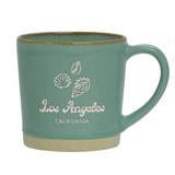 The Los Angeles Shells Etched Ceramic Mug is a 12 oz light green mug with a beige base, decorated with white seashell illustrations and "Los Angeles CALIFORNIA" printed on the side.