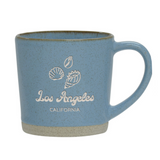 The Los Angeles Shells Etched Ceramic Mug is a 12 oz light blue mug with a speckled texture, white seashell illustrations, and "Los Angeles CALIFORNIA" text on the front. Its unglazed base shows the natural clay color.
