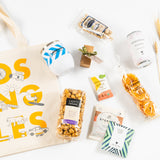 The Los Angeles Wine & Snack Tote features a custom logo tumbler with California snacks, caramel popcorn, dog treats, chocolate, cookies, an apple fruit snack, a cider packet, and wheat stalks—all arranged on a white background.