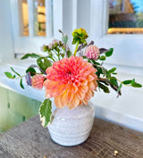 The Love Bud Vase, a small white speckled vase, displays a peach-pink dahlia, clover flowers, and greenery on a wooden surface before a white window frame for a romantic touch.