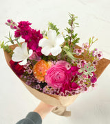 A hand holds the Market Blooms Bouquet—designer’s choice mixed, sustainably grown flowers like pink and orange ranunculus, white cosmos, magenta stock, and pink waxflowers—wrapped in brown paper against a light background.
