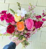 A hand holds the Market Blooms Bouquet—fresh seasonal flowers in pink, red, yellow, and white with greenery and small pink blossoms—set against a pale green shiplap background.
