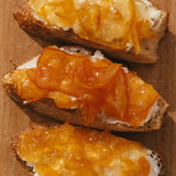 Three slices of bread topped with creamy cheese and Ruby Valencia Orange and Apricot Marmalade on a wooden surface.