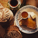 A plate with buttered bread and Sunrise Pixie Tangerine Marmalade, a loaf, coffee, and butter on a wooden table.