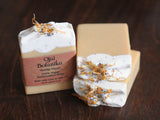 Two Matilija Poppy Ojai Botanika Soap bars with creamy white tops, dried flower petals, black poppy seeds, and organic calendula sit on a dark wooden surface. One has a brown label reading “Ojai Botanika Matilija Poppy Handcrafted Soap.”.