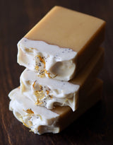 Three Matilija Poppy Ojai Botanika Soap bars stacked on a dark wooden surface, each topped with a creamy white layer and decorated with organic calendula petals and seeds. Infused with lemongrass essential oil for a refreshing, natural scent.
