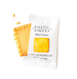 A Meyer Lemon Shortbread Cookie in white packaging sits beside an unwrapped, scalloped-edged square of the same cookie with a few crumbs on a white background.