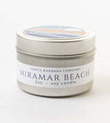 A 3oz silver tin Miramar Beach Travel Candle by Santa Barbara Company, featuring a coastal illustration on the lid and an ocean breeze scent.