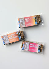 Three Ocean Ranch Handcrafted Granola Bars sit on a white surface in Coconut Almond Chocolate, Maple Chai, and Vanilla Raspberry flavors. Each wheat-free bar comes in a colorful wrapper.