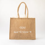 The Ojai Coordinates Jute Tote is a tan, eco-friendly bag with two handles, white tissue paper inside, and "OJAI 34.45° N / 119.24° W" printed in white on the front, shown against a plain white background.