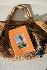 A woven straw bag with an Ojai Guidebook and sunglasses on top, in bright natural light.