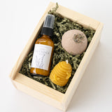 The Ojai Mini Spa Gift Box includes wellness essentials: Ojai lavender mist, a bath bomb with dried lavender, and a beeswax beehive candle, all in a wooden box with shredded green paper.