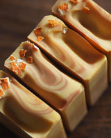 Four Ojai Pixie Handcrafted Soap bars, featuring citrus scent, beige and brown swirls, and dried orange peel topping, are arranged neatly on a dark wooden surface for a refreshing touch.