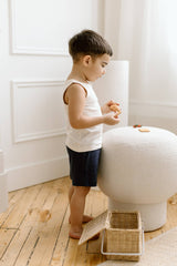 Young boy in the Organic Baby and Kids Leni Tank stands indoors, holding cookies by a round ottoman and a wicker basket.