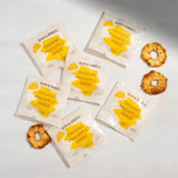 Six Sun & Swell Organic Dried Pineapple snack packs are displayed on a light surface with four dried pineapple rings. The yellow and white compostable packaging makes this an eco-friendly snack choice featuring clear labeling.