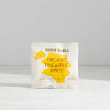 A white pouch of Sun & Swell Organic Dried Pineapple with compostable packaging stands on a white surface against a light gray background. Yellow geometric shapes and text highlight its organic, eco-friendly snack qualities.