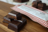 Close-up of stacked Pecan & Medjool Date Dark Chocolate Bar pieces on a wooden surface, revealing their textured filling. An organic chocolate bar package with a red and white checkered design sits in the background, enhancing this tempting vegan dessert scene.
