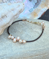 The Pink Pearl Cluster Reflection Bracelet features pink pearls strung on brown leather, shown with seashells in the background.