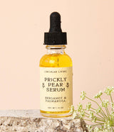 A bottle of Prickly Pear Facial Serum rests on stone, with white flowers in the foreground.