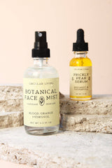 Two skincare bottles on stone slabs: an organic Botanical Face Mist and Prickly Pear Facial Serum with beige labels.