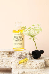 A bottle of Prickly Pear Facial Serum with a dropper and white flower, set on stone blocks against a beige background.