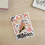 The Quail of California Thank You Note Card is a vibrant recycled paper card featuring an illustrated bird amid orange, pink, and brown florals with “Thank you” in bold at the bottom, elegantly placed on a wooden surface.