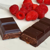 Two pieces of Raspberry & Medjool Date Dark Chocolate Bar, one broken to show its filling, rest on a white surface with a pile of fresh raspberries in the background.