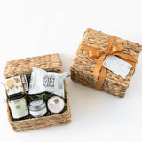 The Renewal Gift Basket features a lavender candle, artisanal soap, bath salts, matches, and a small jar in a woven basket with a gold ribbon, displayed open beside its lid and another closed ribbon-tied basket.