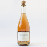A bottle of Riverbench Brut Rosé sparkling wine from Santa Maria Bench, topped with a cork and wire cage, with a white and gold label, set against a plain white background.