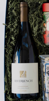 The Riverbench Moments Gift Box features a bottle of Chardonnay from Santa Barbara, nestled in green crinkled paper with other partially visible gifts.