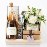 A gift box containing Riverbench Brut Rosé champagne, a selection of Dick Taylor chocolate bars including Black Fig Fine Drinking Chocolate and Local Trio of Caramels, and an assortment of gourmet chocolate covered candies. The box is accompanied by a fresh floral arrangement.