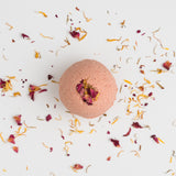 The Riviera Gardens Bath Bomb, a pink sphere with rose kaolin clay and dried flower petals at its center, rests on a white surface amid scattered yellow and red dried petals.
