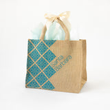 The Riviera Wine Tote is a jute bag with blue geometric patterns and “Santa Barbara” lettering, filled with white tissue and finished with a cream bow on the handles—perfect for Santa Barbara gifts, shown on a white background.