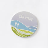 The San Diego Vistas Coaster showcases hills, two palm trees, a bird, and “SAN DIEGO” on a pastel sky—an ideal scenic ceramic stone coaster for fans of San Diego.