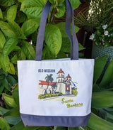 The Santa Barbara Mission Canvas Tote is a white USA-made bag with dark handles, screen-printed with the Old Mission, palm trees, and “Santa Barbara” in yellow. It's shown amid lush green plants.