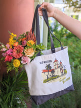 A hand holds the Santa Barbara Mission Canvas Tote, screen-printed with the iconic Old Mission. Made in the USA, it's filled with colorful roses and zinnias, all set against a backdrop of lush greenery.