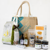 The Santa Barbara Picnic Gift Tote includes artisan popcorn, snacks, nuts, breadsticks, jams, olive oil, and more gourmet treats in a stylish gift bag, all arranged neatly against a white background.