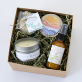The Santa Barbara Spa Petite Gift Box includes a candle, soap bar, round tin with seaside bath bomb in a colorful sunset design, and Citrus Sunshine Mist, all arranged on green shredded paper.
