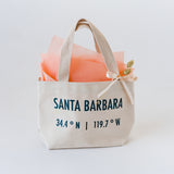 A Santa Barbara Sunset Traveler Tote with “Santa Barbara 34.4° N | 119.7° W” in dark blue, filled with peach tissue, a bow, dried wheat stalk, and space to add a plant-based soy wax Santa Barbara travel candle.