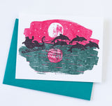 The Santa's Dolphins Letterpress Holiday Card Boxed Set features a holiday card with Santa’s sleigh pulled by dolphins over ocean waves beneath a full moon and pink sky, paired with a teal envelope.