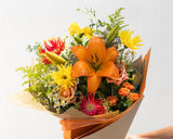 A vibrant mixed flower bouquet wrapped in brown kraft paper, held by a person's hand against a bright white background.