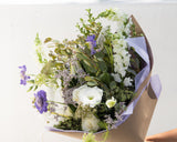 Hand-Tied Bouquet: The Lush featuring white roses, snapdragons, and purple scabiosa wrapped in brown and lilac paper.