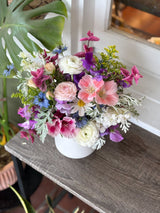 The Full: Seasonal Flowers Arrangement in a white vase is displayed on a wooden table by a window, framed by green foliage and featuring vibrant pink, white, purple, and blue seasonal blooms.