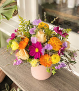 The Full Seasonal Flowers Arrangement features orange, yellow, purple, and cream blooms in a pastel pink pot, set on a wooden surface by a sunlit window with green foliage in the background.