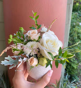 A hand holds the "Seasonal Flowers - Cup" featuring white and pale peach roses, greenery, and delicate filler flowers, set against an outdoor backdrop with a pink column and lush greenery. Local Santa Barbara delivery only.