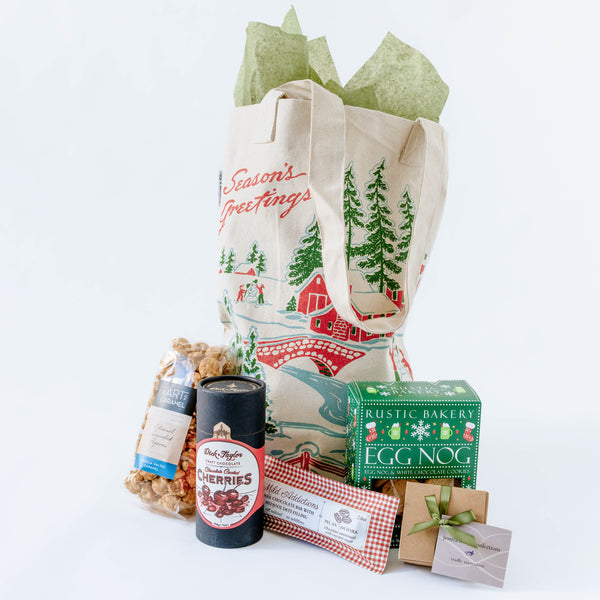 Holiday gift basket with a tote bag, eggnog cookies, chocolate cherries, and other items on a white background
