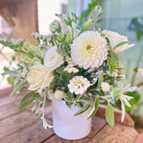 The Serene Mama Arrangement features elegant white roses and dahlias with green foliage in a white ceramic vase, all sourced from local farm flowers, beautifully displayed on a wooden surface in soft natural lighting.