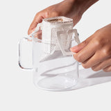 A person places a Single-Serve Pour Over Churro Coffee drip filter bag over the rim of a clear glass mug, preparing to brew delicious coffee.